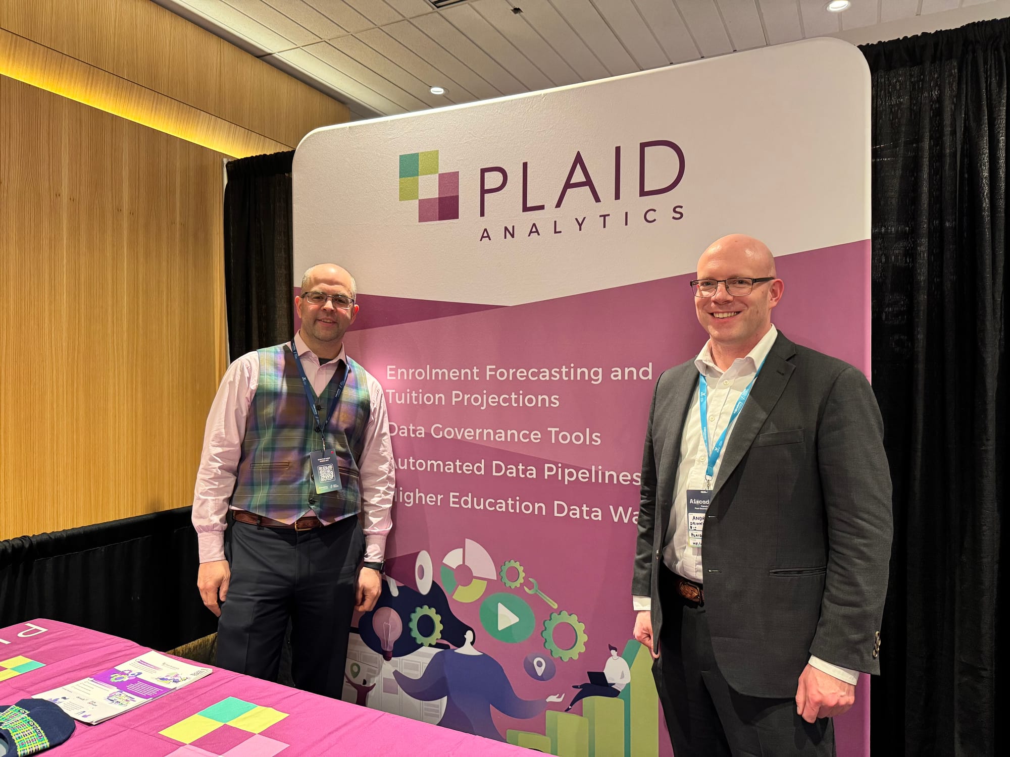Andrew and Pat at the AI-Cademy Conference in front of purple and white Plaid Analytics banner outlining enrolment and tuition forecasts, data governance tools, automated data pipelines, and data warehouses