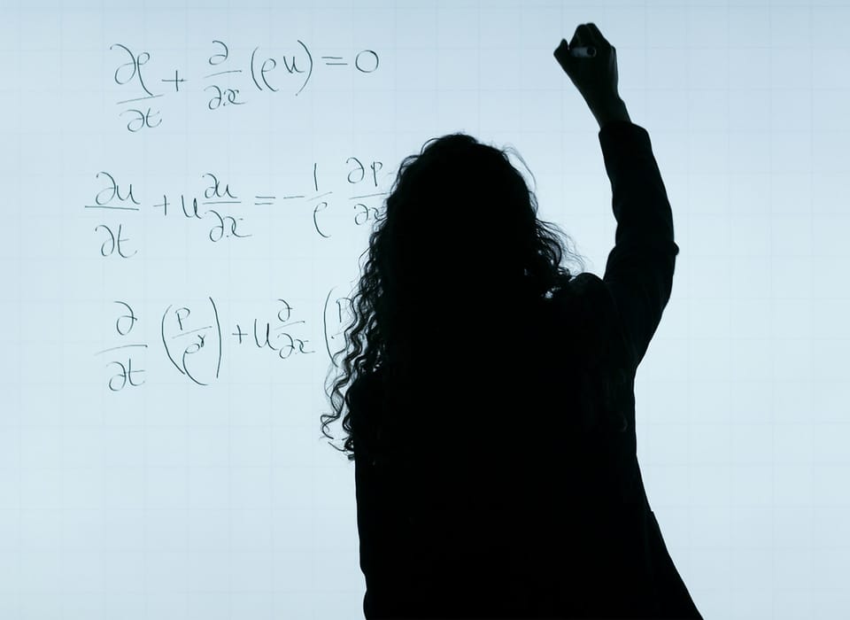 A woman is silhoutted against a white board while she writes out a complex algebraic formula.